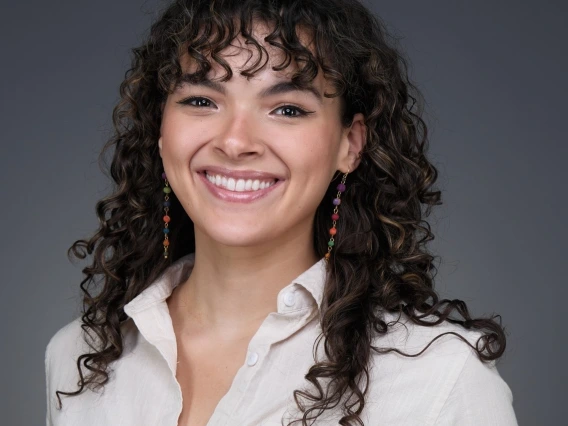 Headshot of woman with long brown curly hair and bangs wearing a white button-down shirt and dangly earrings smiling at the camera