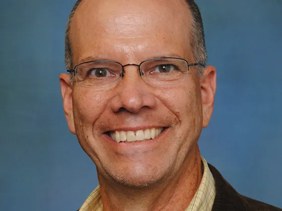 Headshot of balding man wearing glasses, yellow shirt and dark blazer smiling at the camera.