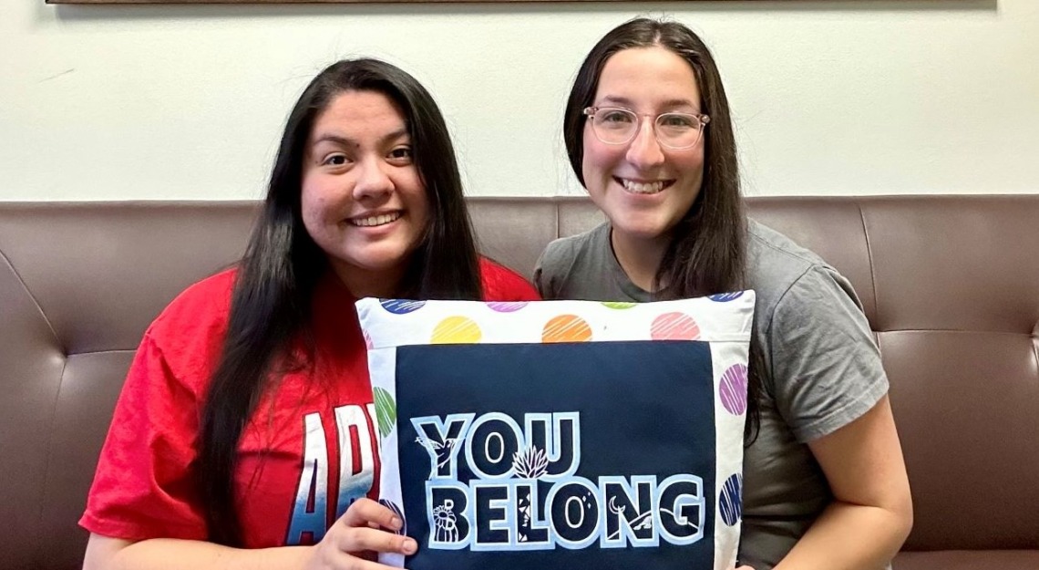 Two undergraduates smiling and holding message pillow You Belong