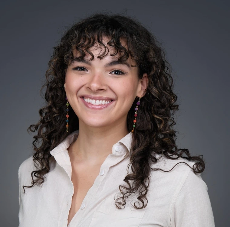 Headshot of woman with long brown curly hair and bangs wearing a white button-down shirt and dangly earrings smiling at the camera