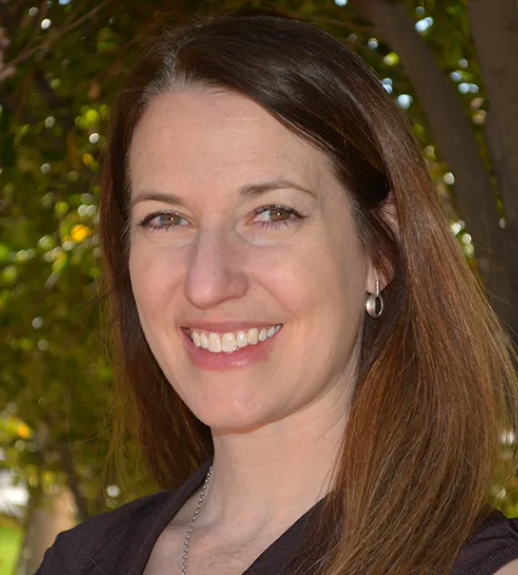 Headshot of woman with medium brown shoulder length hair parted on the side wearing a dark shirt smiling at the camera.