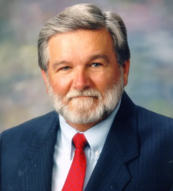 Headshot of man with gray hair, beard, and mustache wearing blue coat and shirt and red tie looking at the camera.