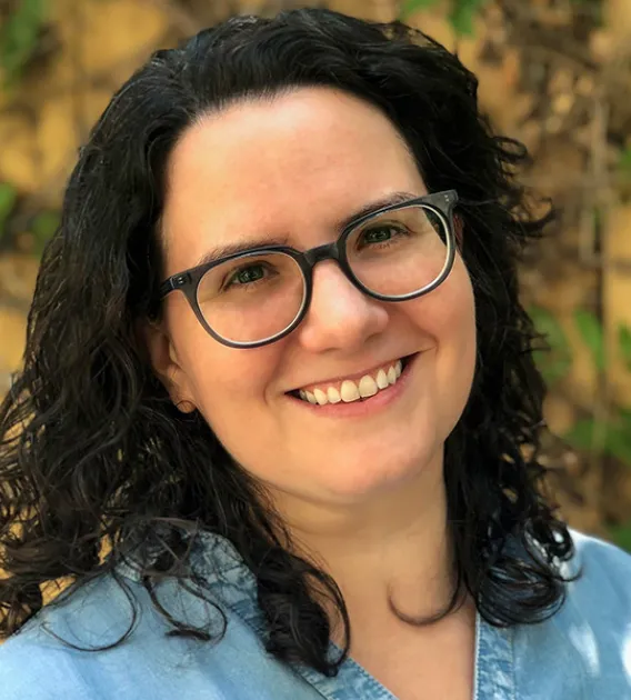 Headshot of woman with dark brown shoulder length hair and wearing glasses and a blue shirt standing in front of yellow wall with foliage smiling at the camera.