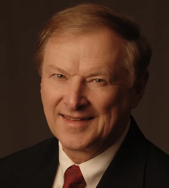 Headshot of older man with light hair wearing yellow shirt, red tie and dark blazer smiling at the camera.