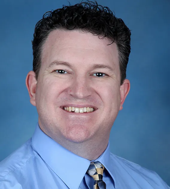 Headshot of man with brown short hair wearing blue shirt and patterned tie smiling at the camera.