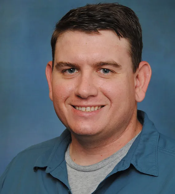 Headshot of man with short brown hair wearing blue collared shirt with gray t shirt underneath smiling at the camera.