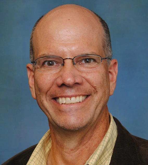 Headshot of balding man wearing glasses, yellow shirt and dark blazer smiling at the camera.